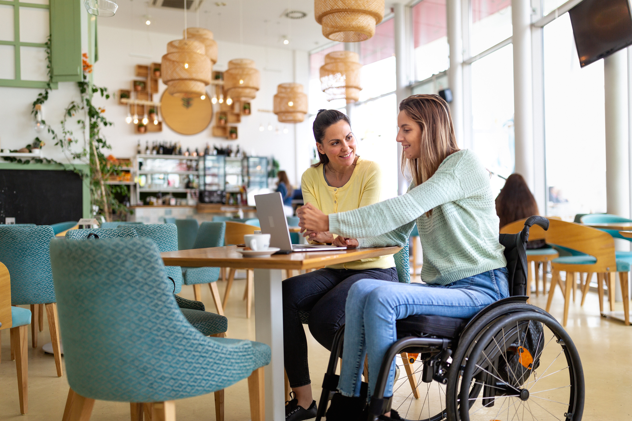 Two people, one in a wheelchair, using a laptop in a cafe.