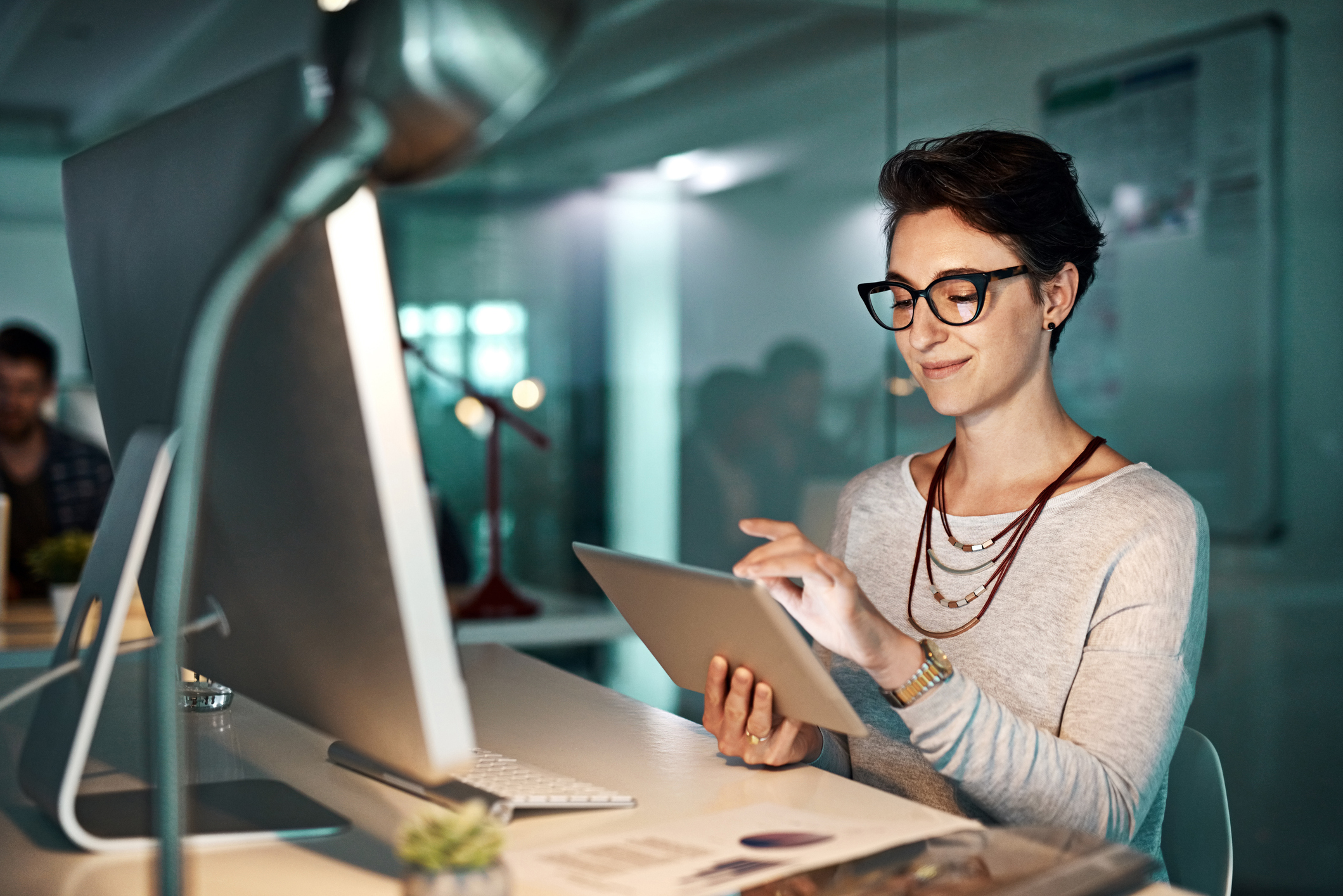 An investor in an office setting sits in front of a computer and uses a tablet.