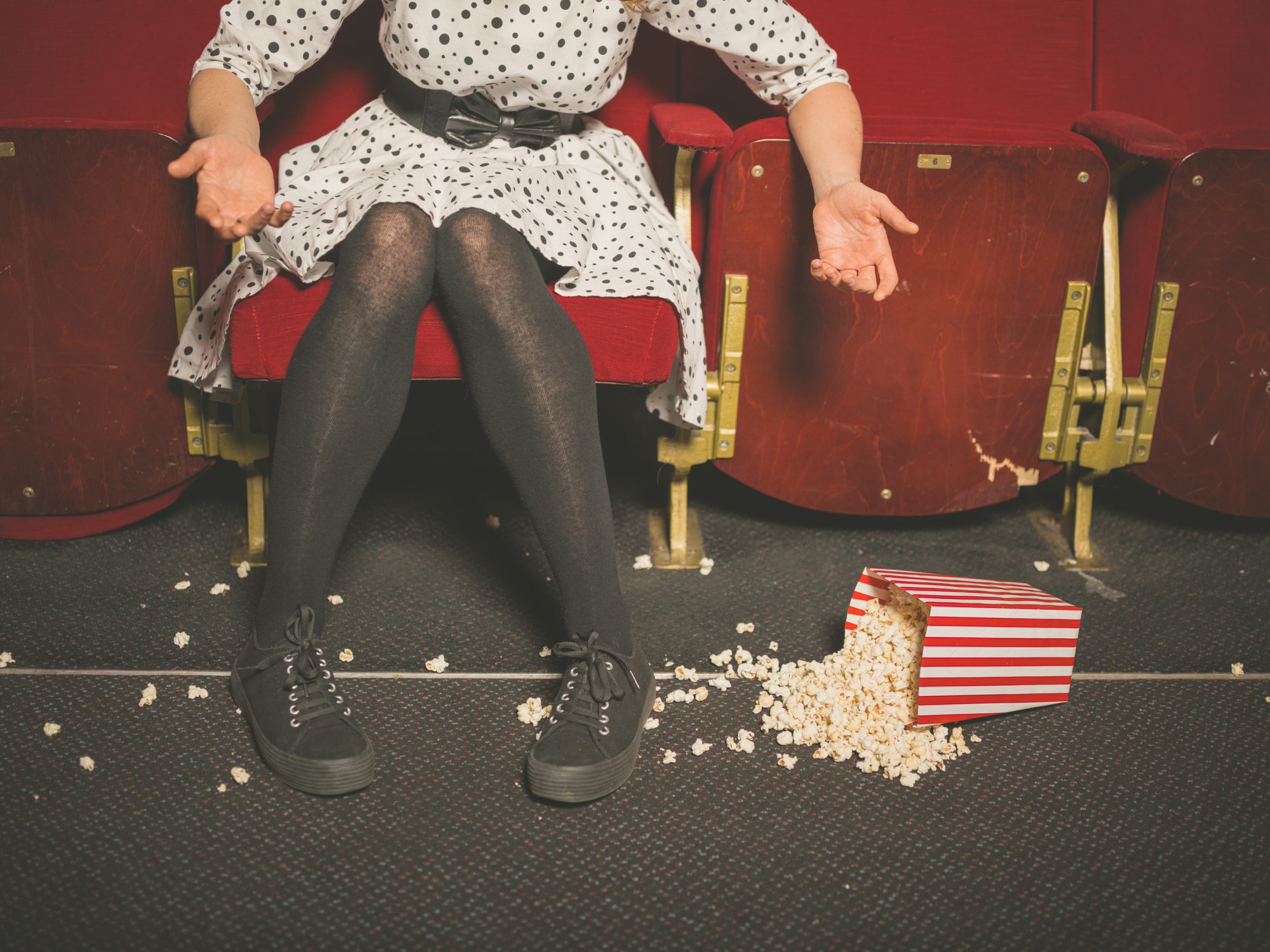 A woman sits in a movie theater seat reacting to a dropped box of popcorn laying on the floor of the theater.