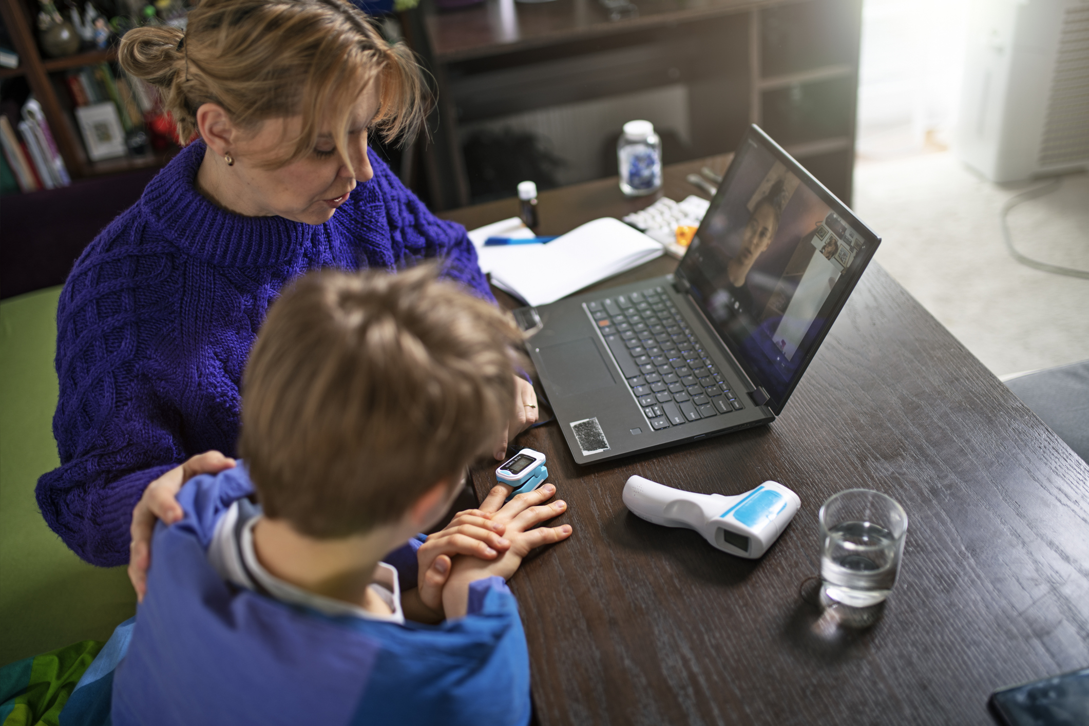 An adult and child attend a telehealth appointment via a laptop, and the child is wearing a pulse oximeter.