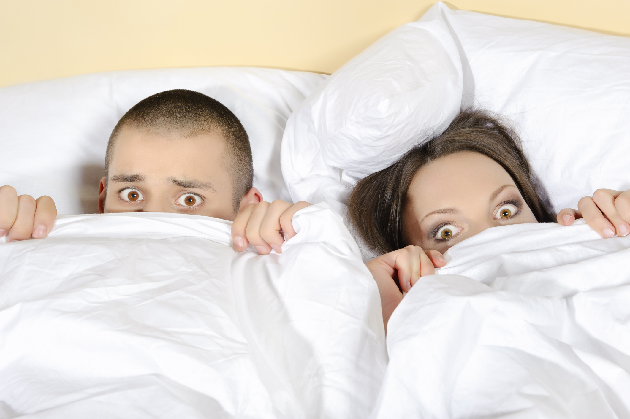 Two people in bed covering the lower halves of their faces with a comforter.
