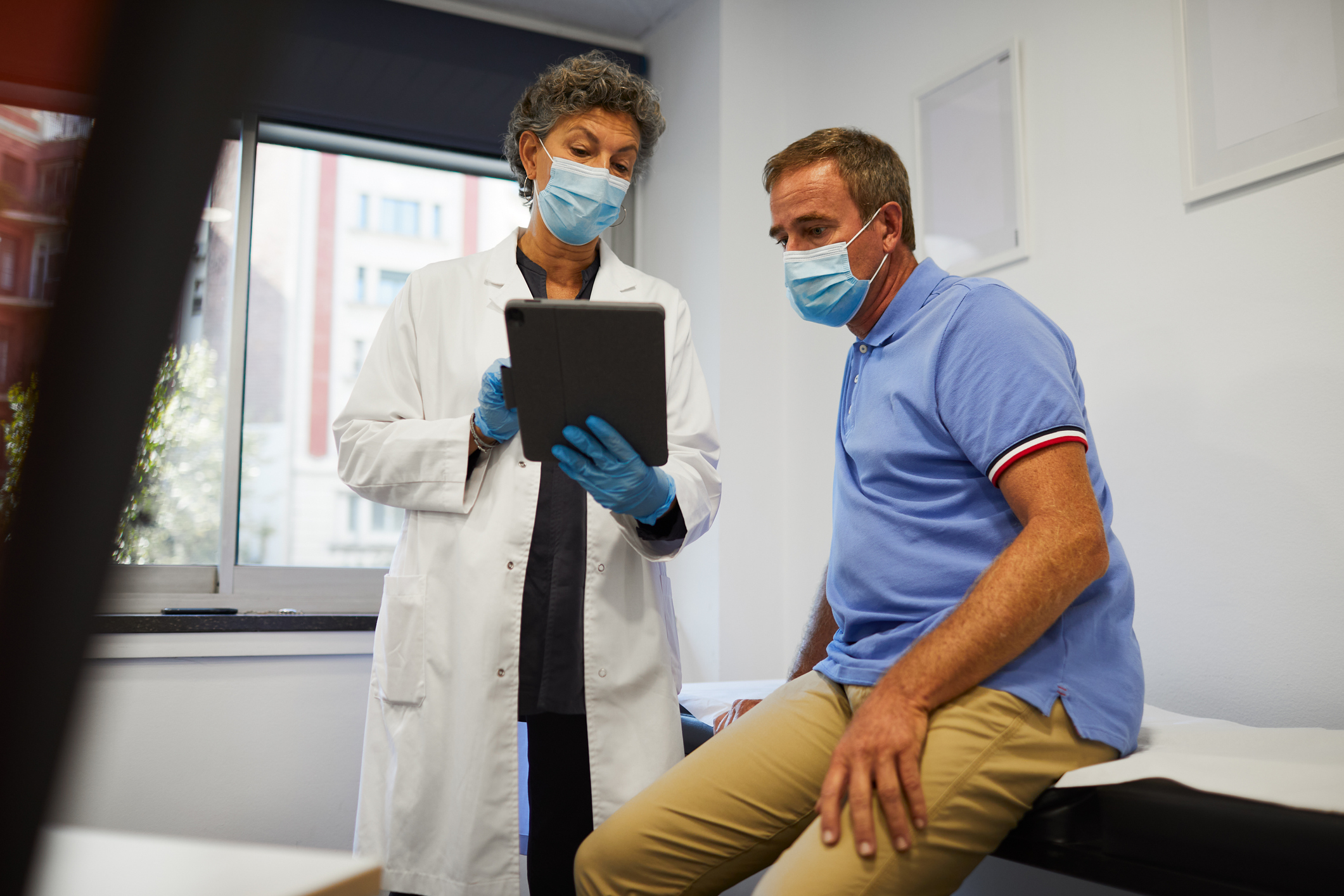 Masked doctor shows a masked patient a page on her iPad.