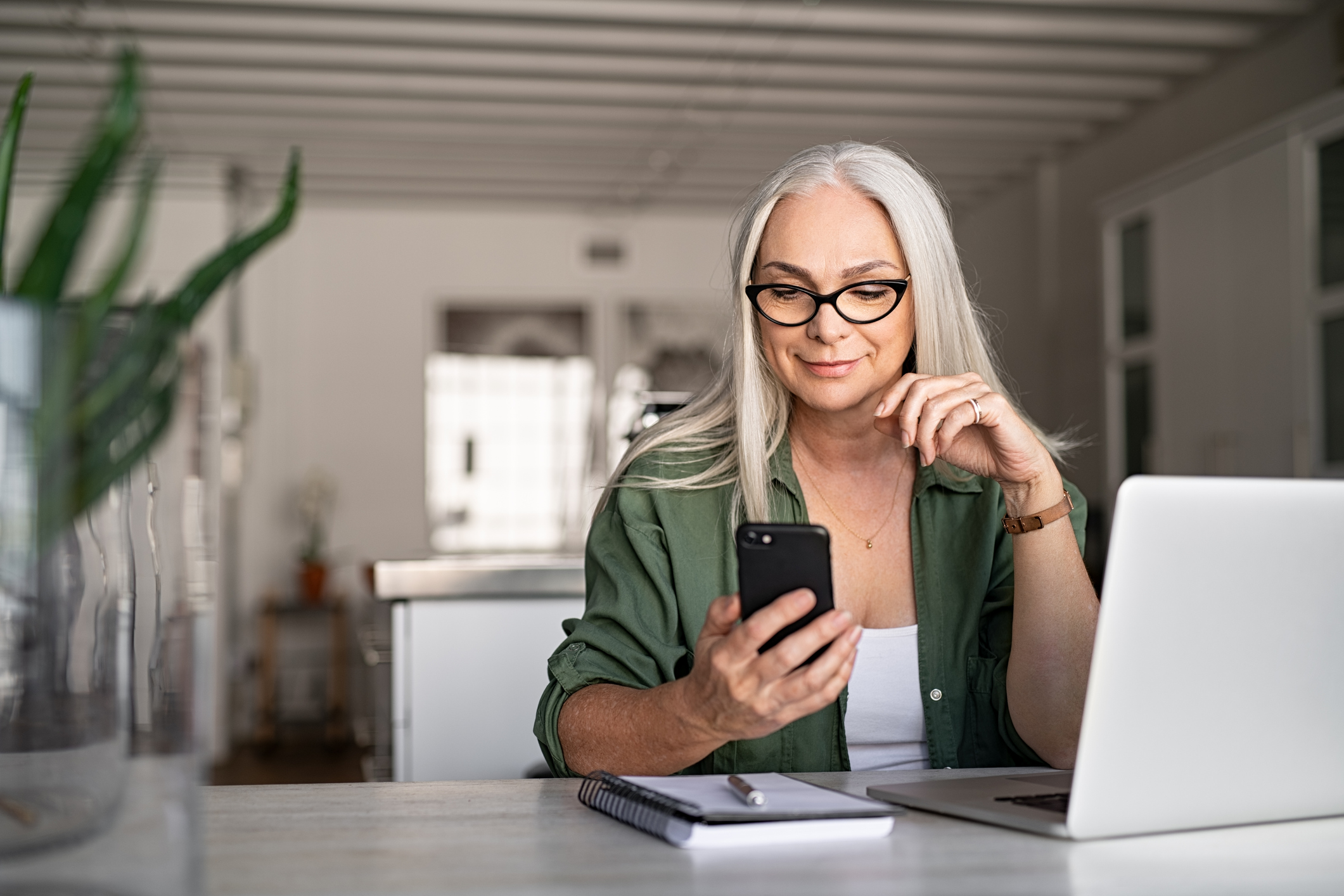 An older woman checks her smartphone while working on a laptop computer.