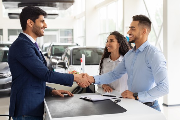 A smiling couple shaking hands with a car dealer inside a dealership after making a purchase.