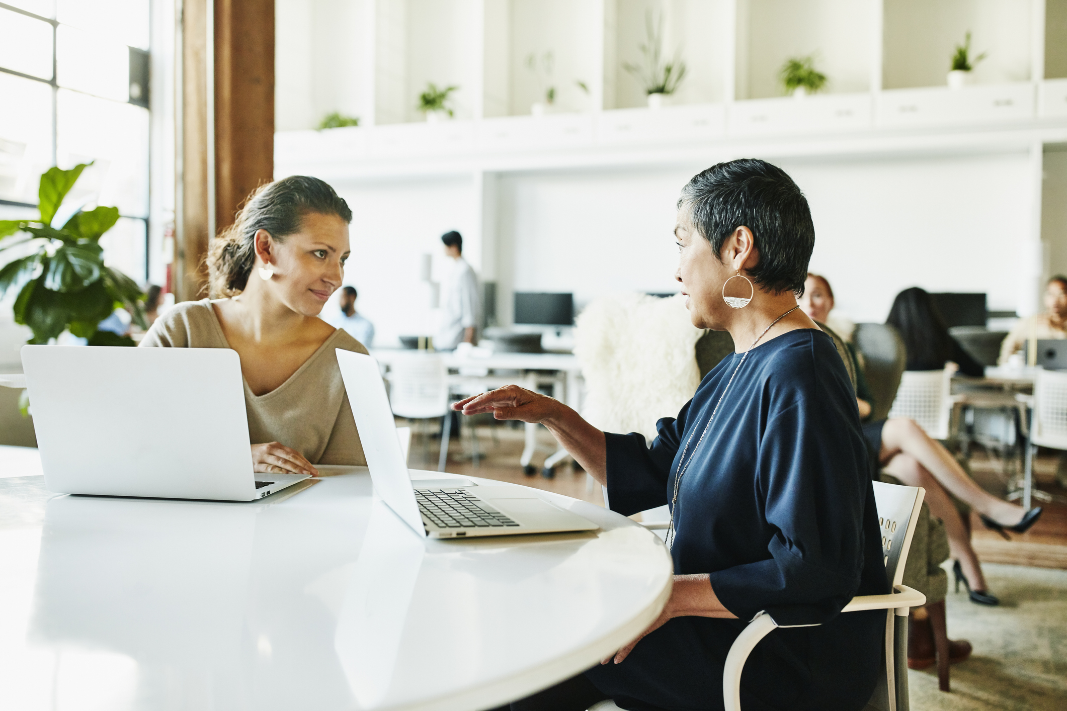Two women at a table sit looking at each other with their laptops open.