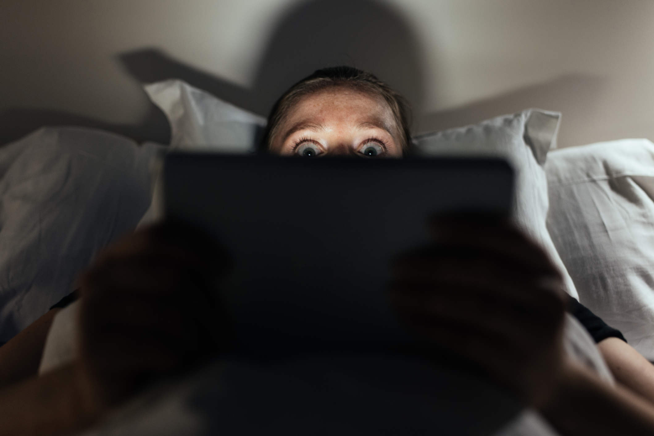 A person with eyebrows raised looking at a touchscreen tablet while lying in bed.
