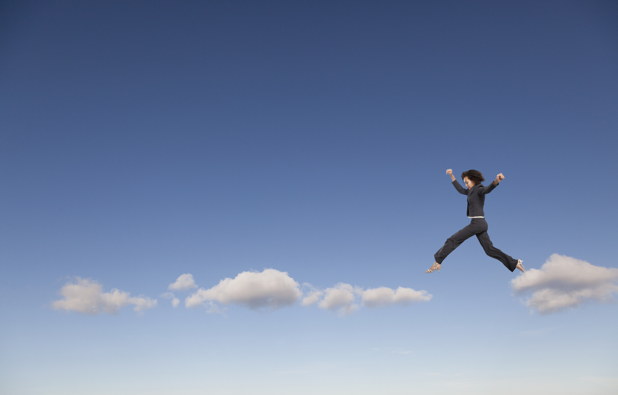 Woman leaps from cloud to cloud in a blue sky.