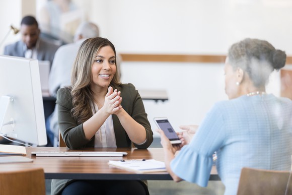 A person looks at their smartphone while sitting at a desk across from another person.