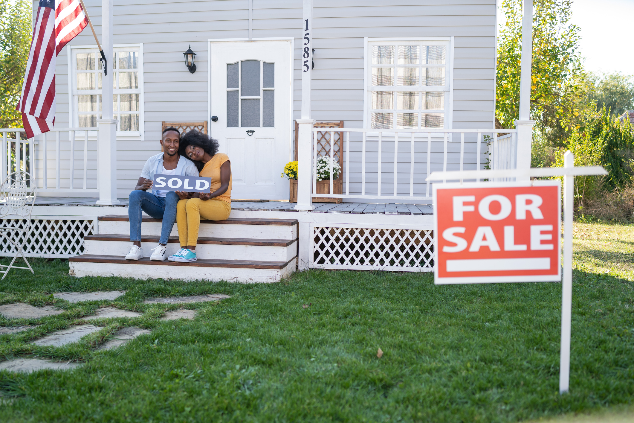Two people sit on the porch of a home with a "For Sale" sign.  They hold a sign that says "Sold."