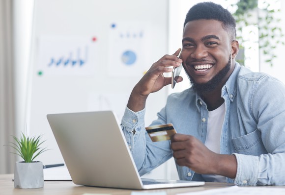 A man holding a credit card in one hand and a mobile phone in the other sitting in front of a computer.
