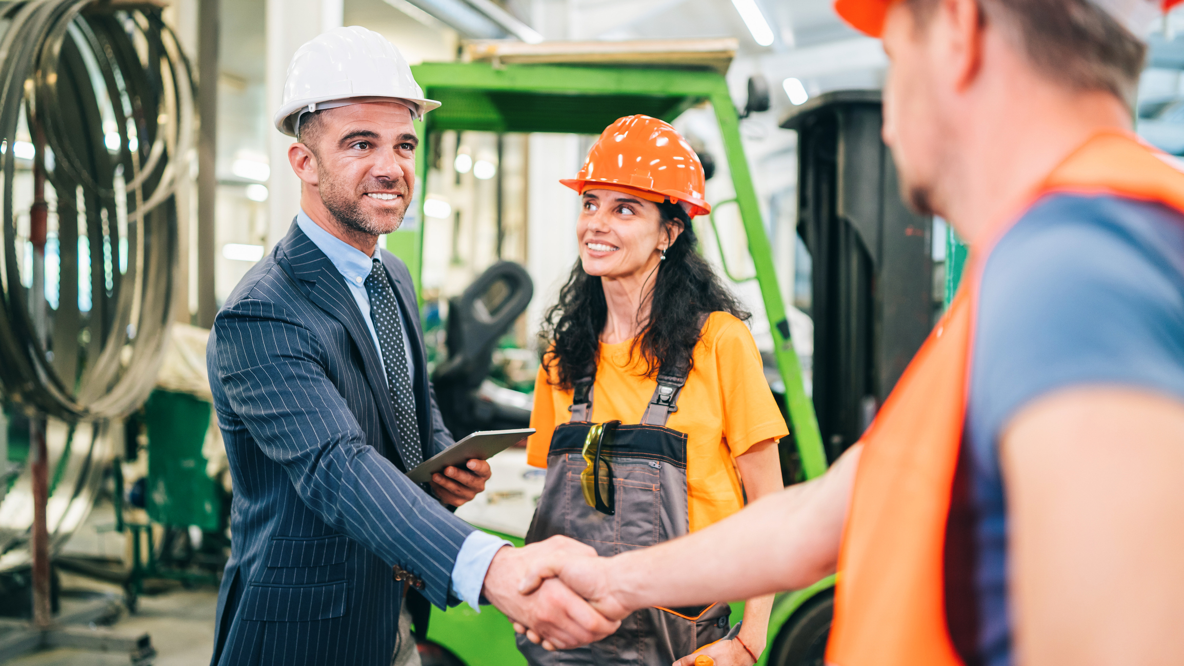A person shaking hands with another in a factory as a co-worker looks on.