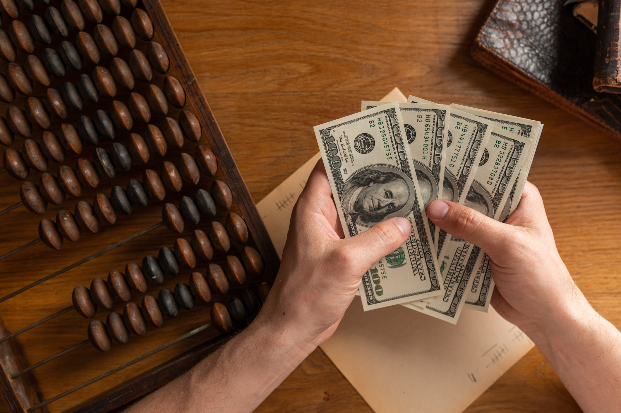 Person holding multiple hundred dollar bills with an abacus next to their hands.