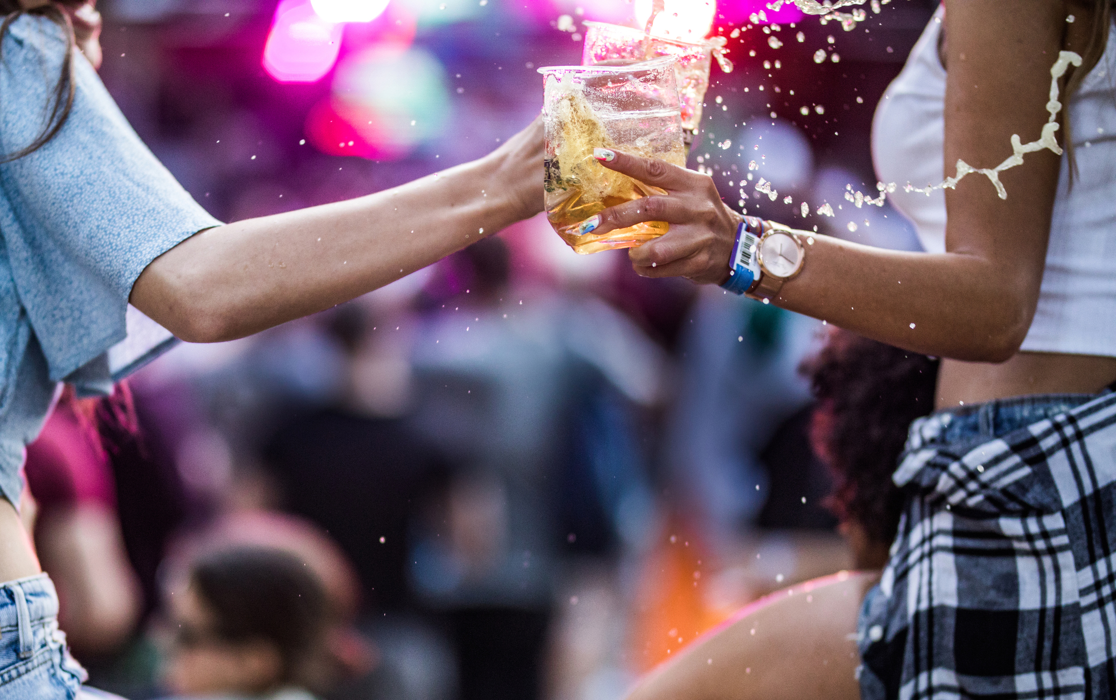 Two people in an outdoor setting push their glasses of alcohol together and spill it everywhere