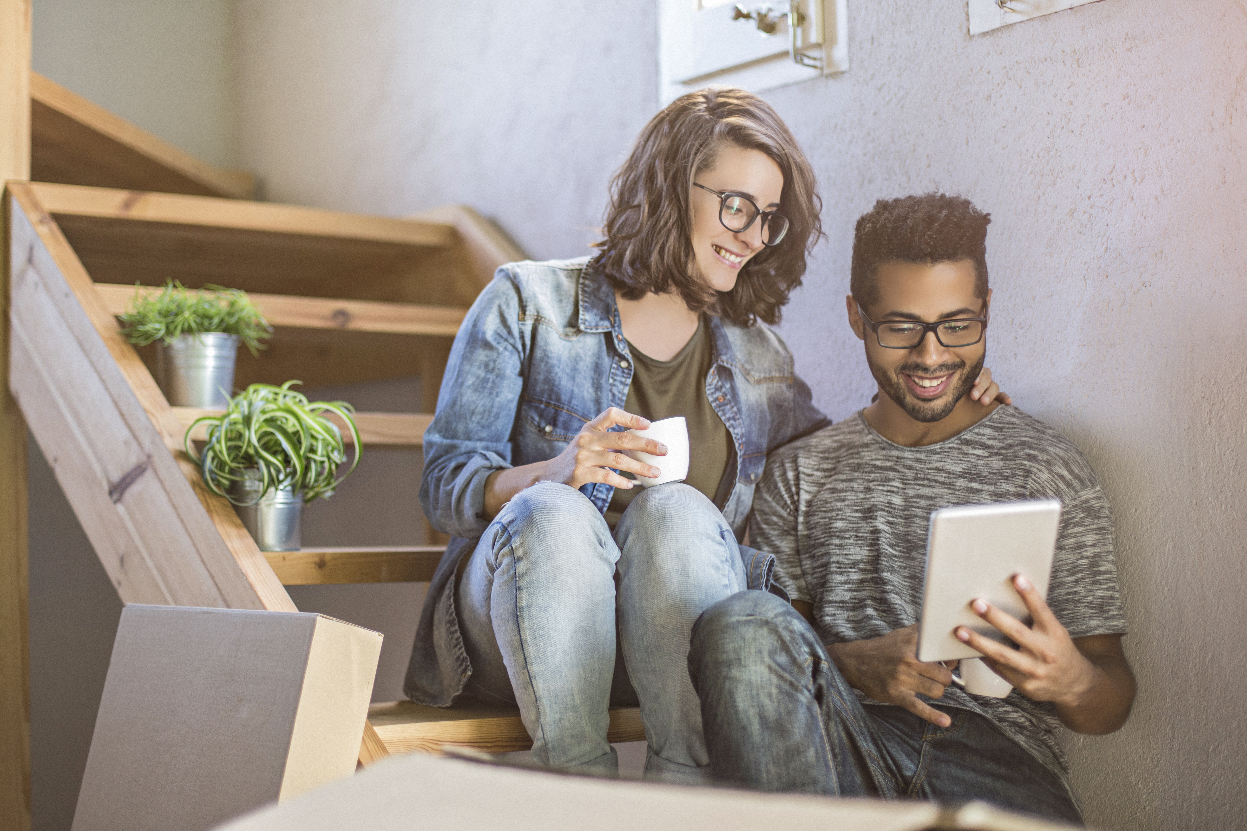 A smiling couple that just moved into a new home, looking at a tablet device while sitting on the stairs.