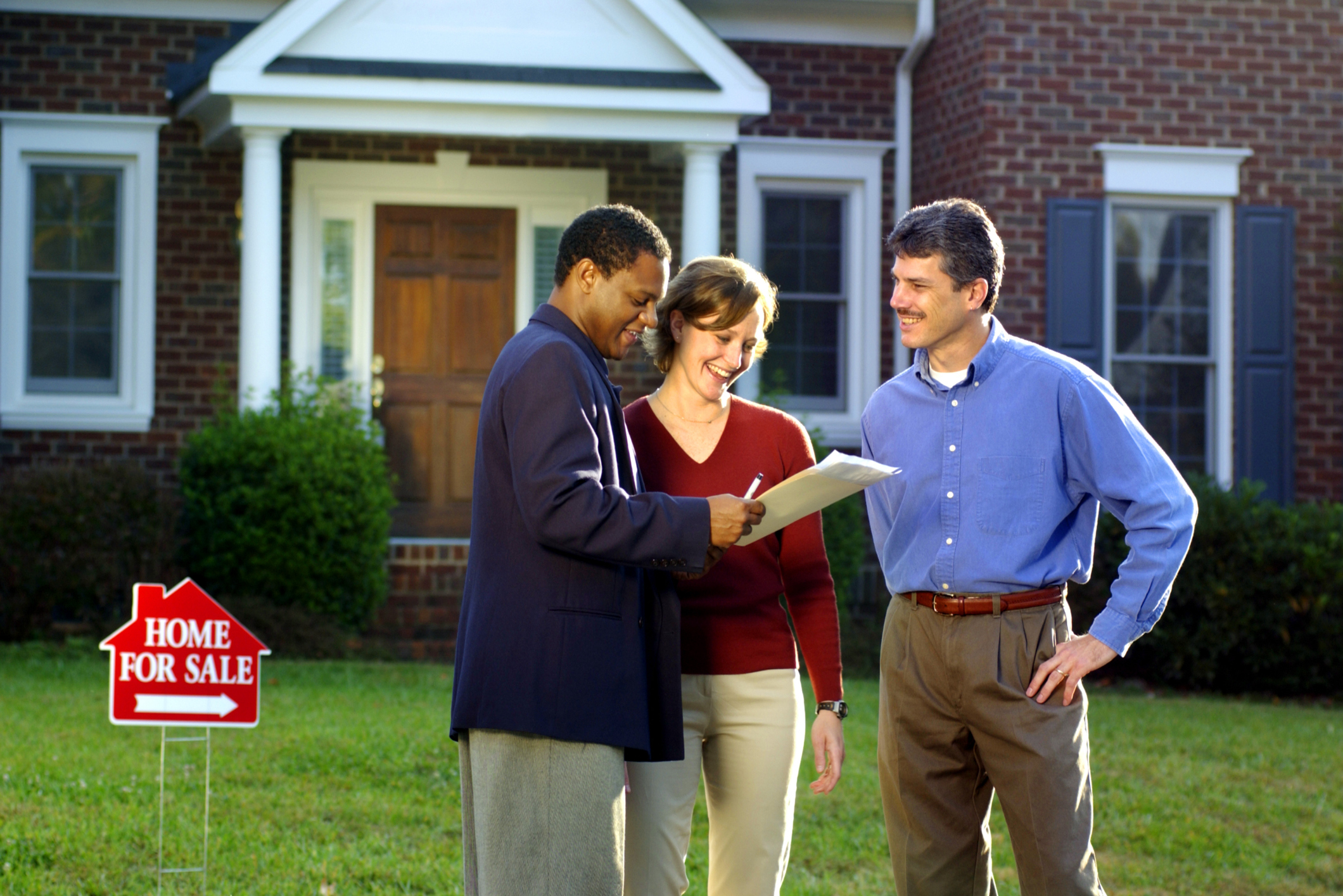 Three people in front of a house with a for sale sign on the lawn.