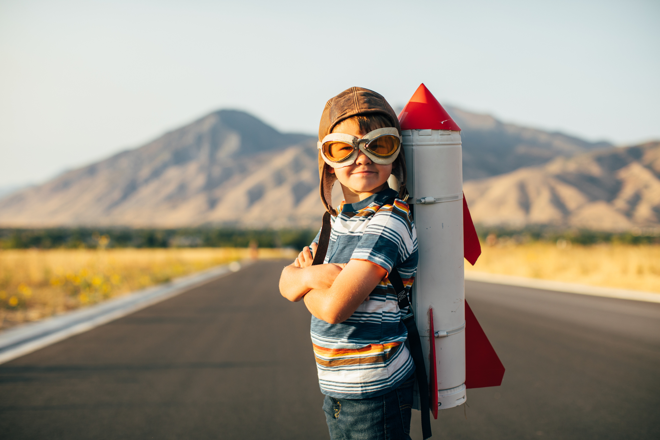 A child wearing aviator goggles and cap with a rocket strapped to their back.