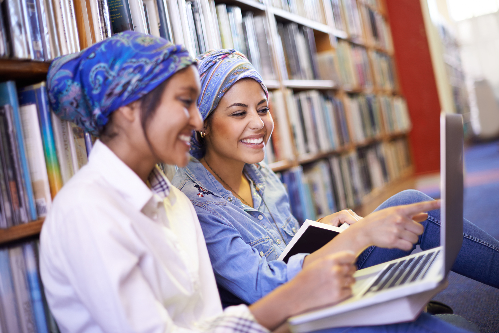 Two smiling people use laptops in a library.