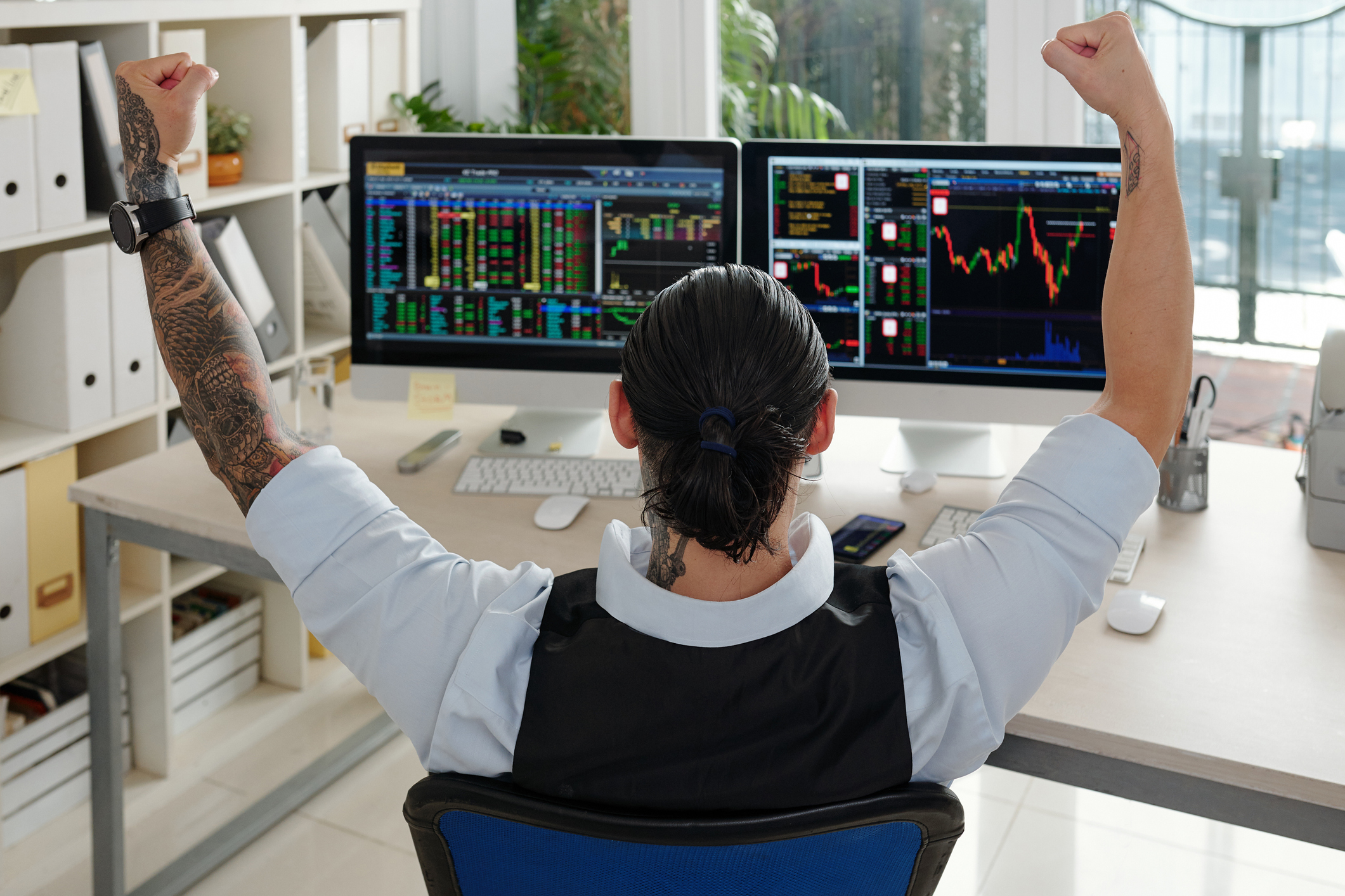 A person raising hands in celebration, while looking at stock price charts on a computer screen.