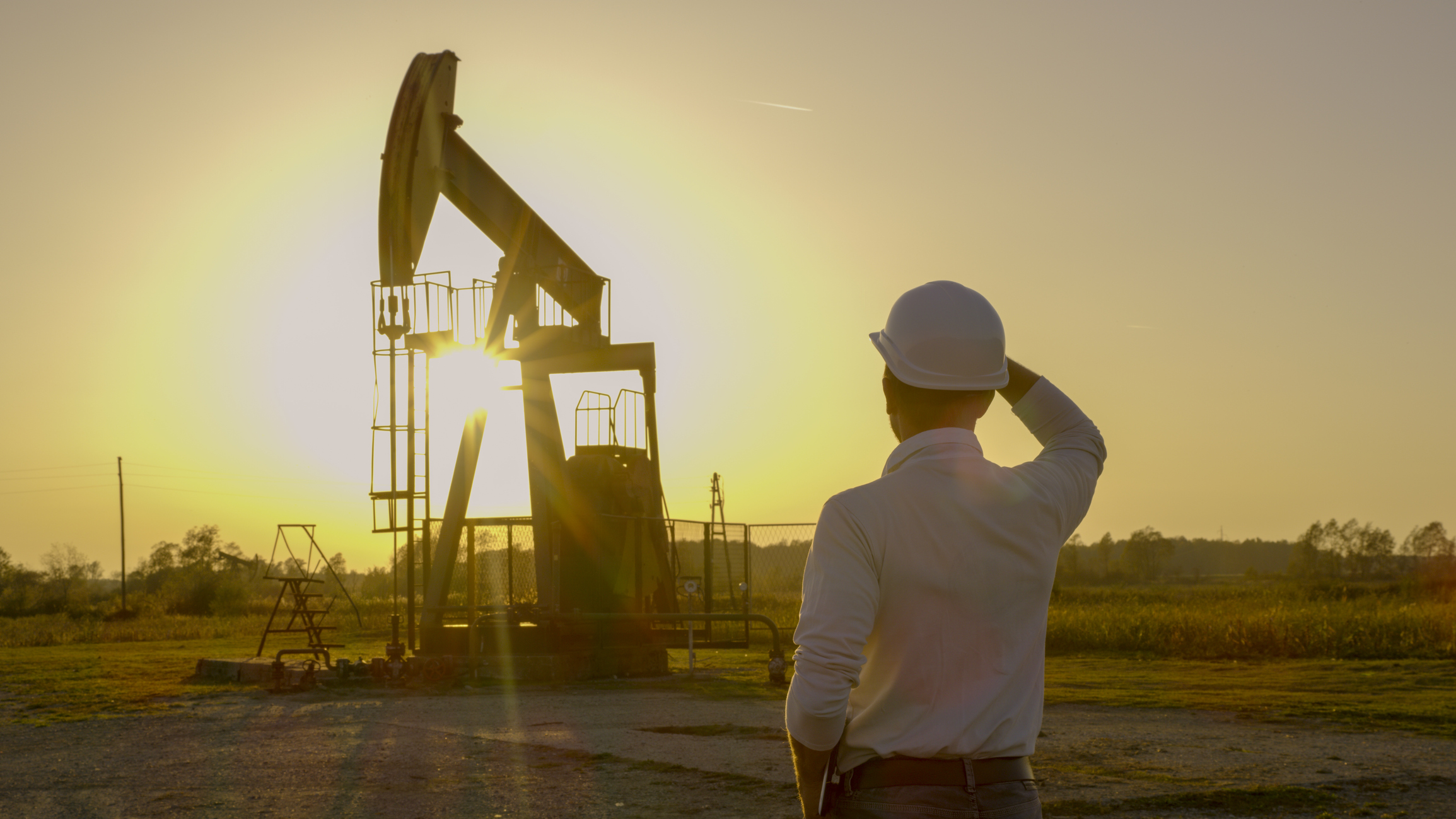 A person wearing a white hard hat looks at a pumpjack in front of a bright full yellow sun.