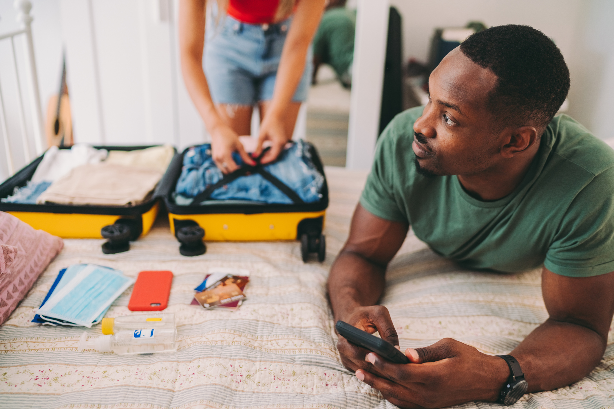 A person lays on a bed while holding a smartphone as another person packs a suitcase with face masks and credit cards.