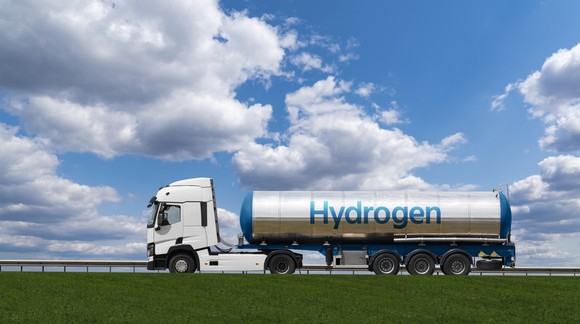 A tanker truck labeled "Hydrogen" drives along a road under a blue sky with clouds.