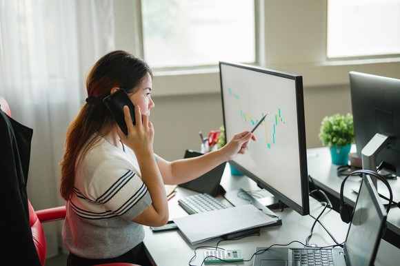 A person sits at a desk on the phone while pointing to a chart on the computer. 
