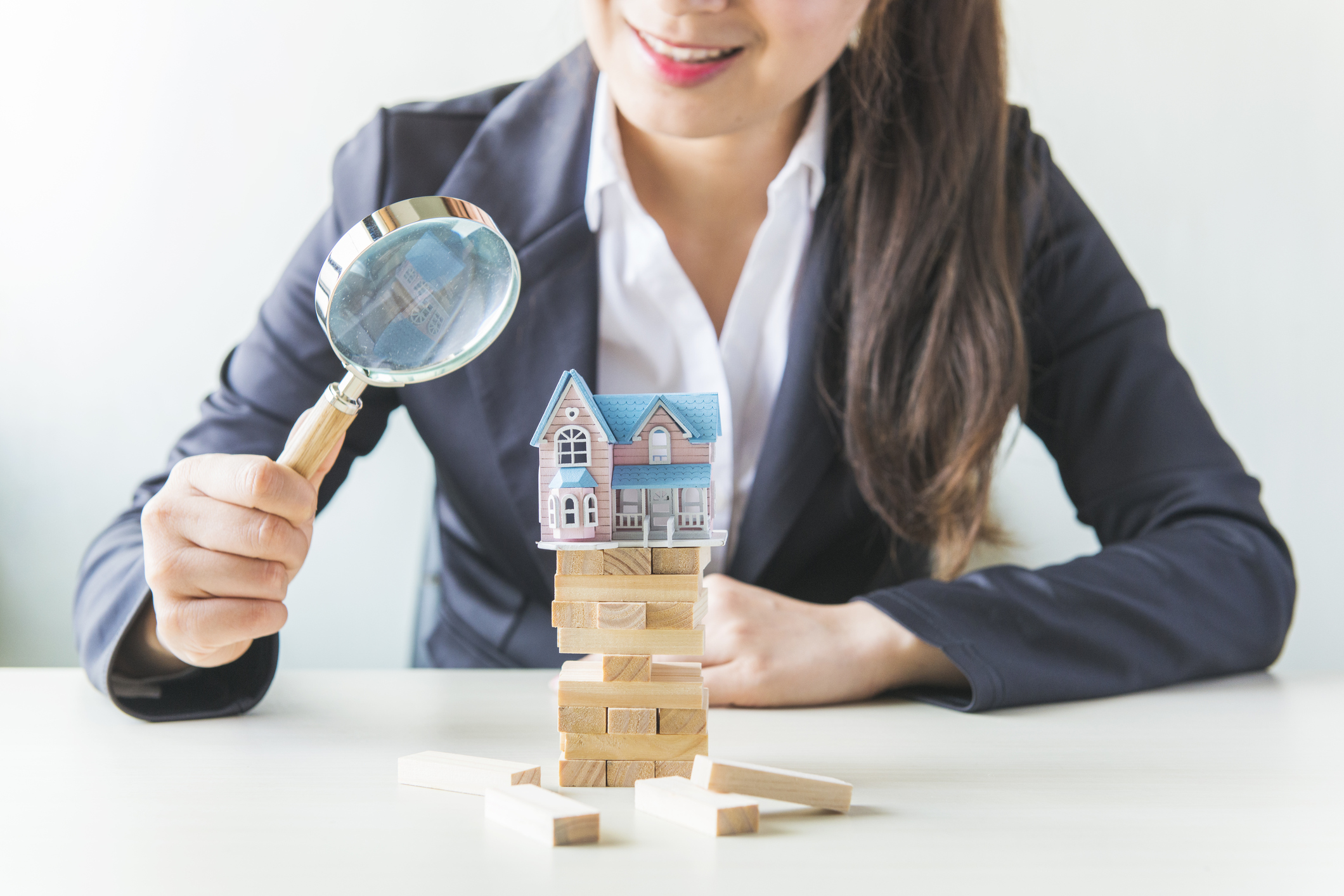 Person with magnifying glass looking at model house on building blocks.
