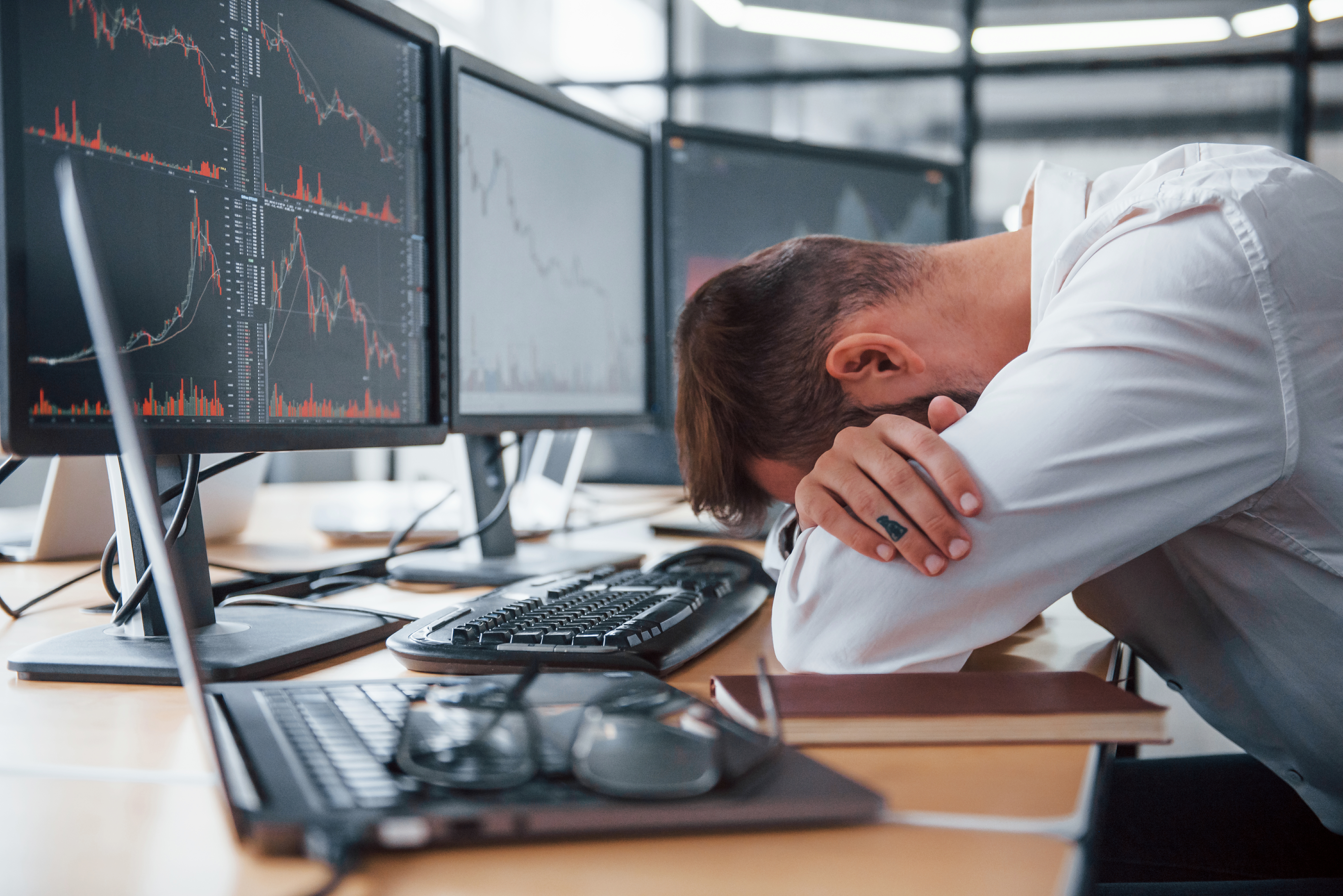 Person with their head resting in their arms at their desk, in front of stock charts on computer monitors.