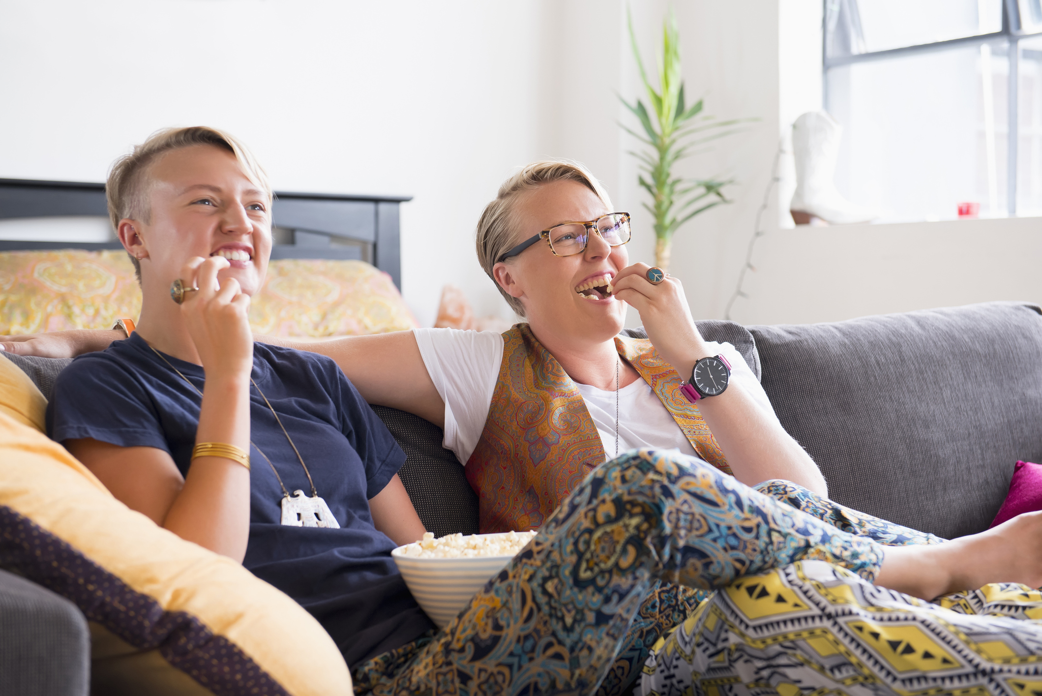 A couple is watching TV, eating popcorn, and smiling while sitting on a couch.