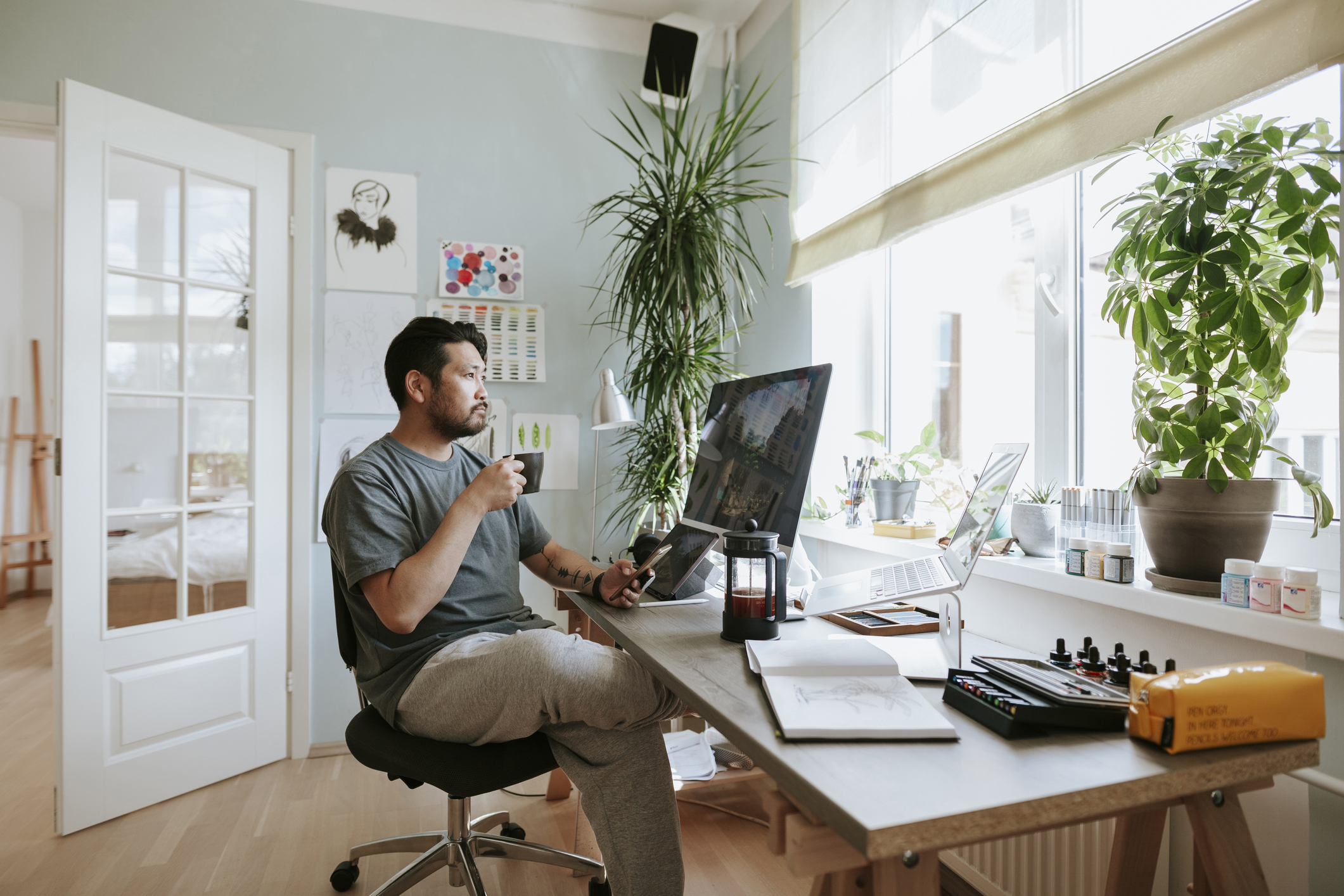 A person holding a drink and working in an open, airy, home office with plants.