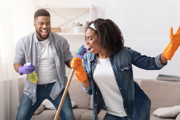 One person holds a spray bottle and smiles while another person holds a broom handle and sings into it like a microphone.