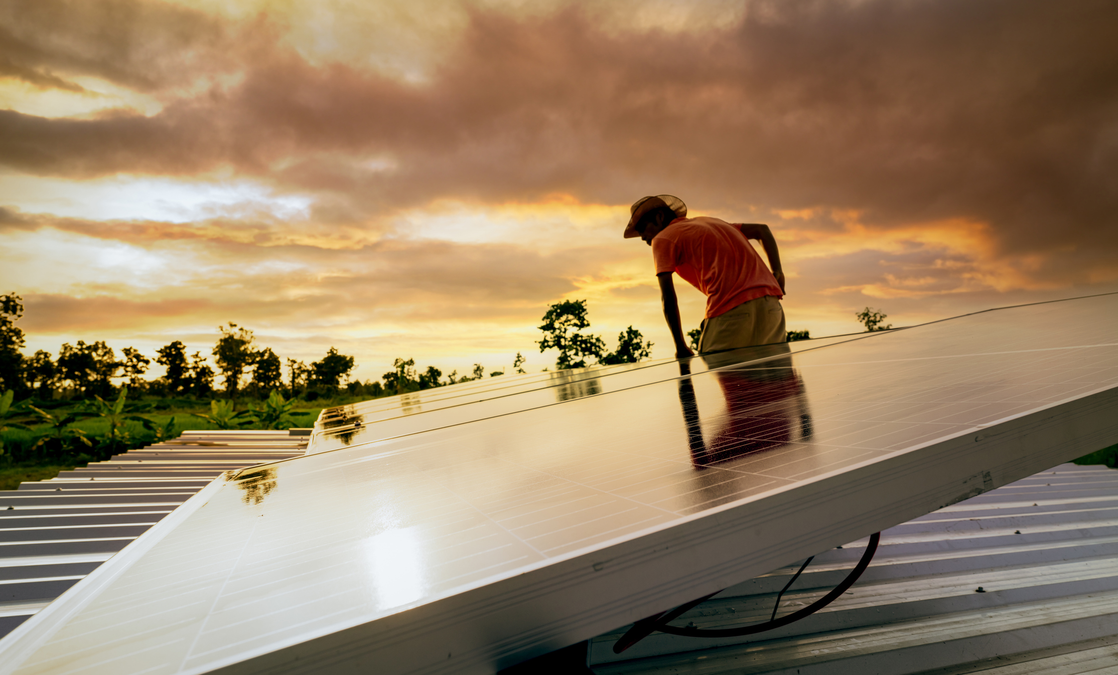 A man inspects a rooftop solar panel with a sunset in the background.