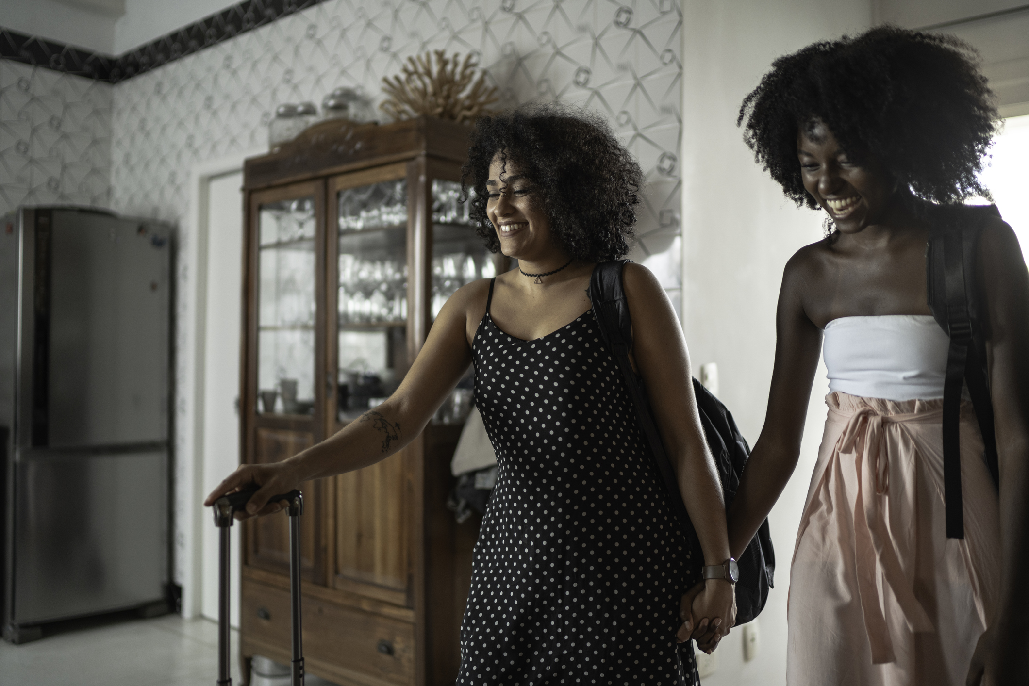 Two people holding hands and carrying luggage into a house.