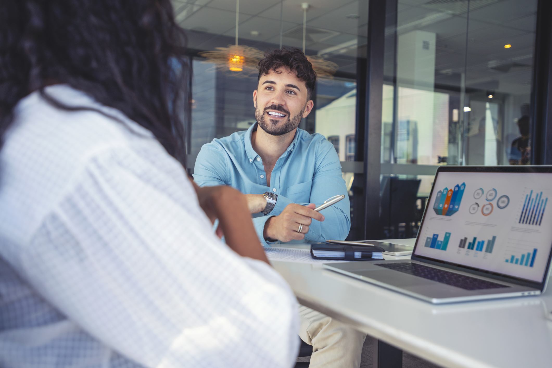 Two people talk in an office, while a laptop displaying charts sits on a nearby desk.