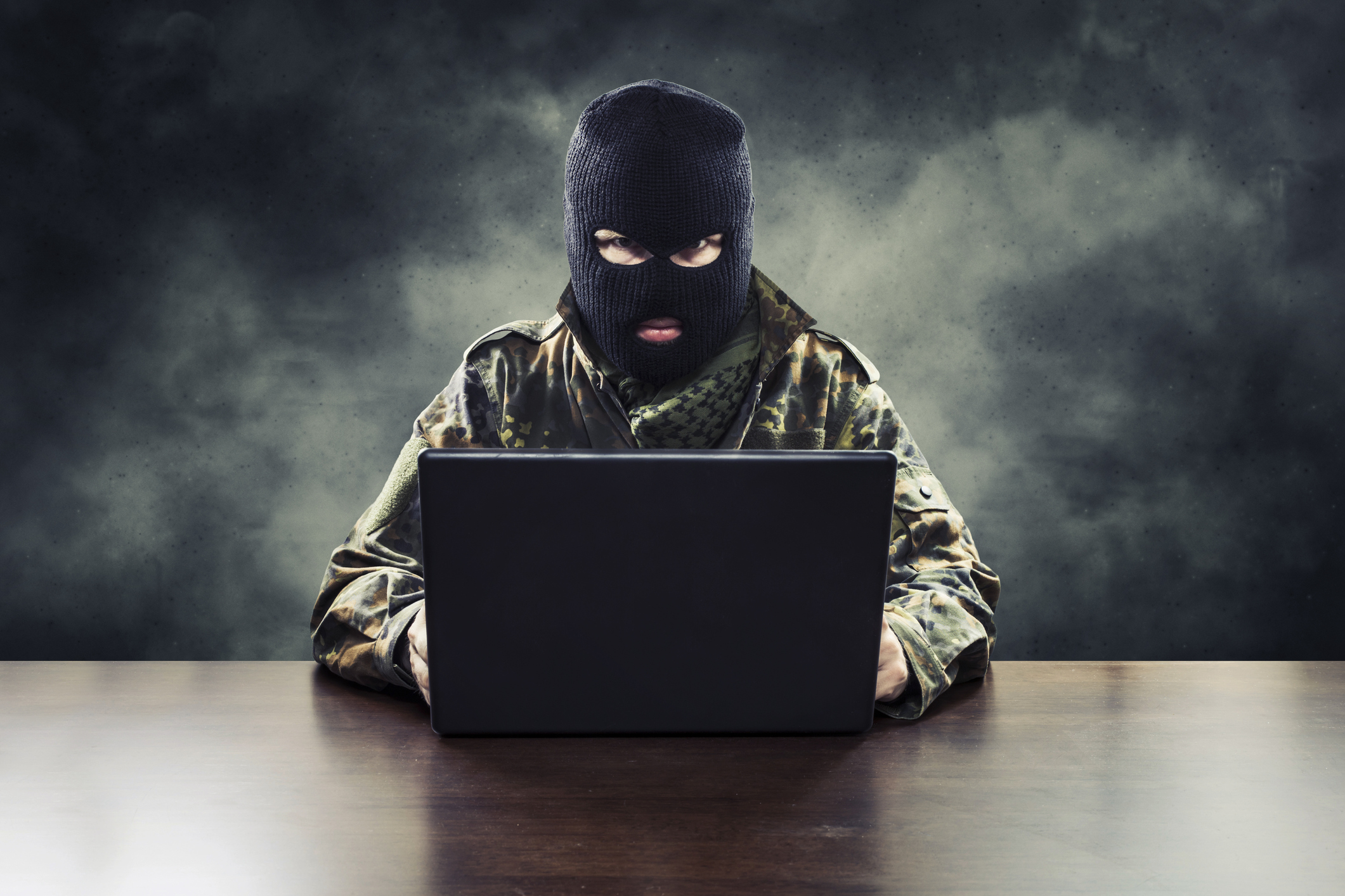 A person wearing camouflage and a ski mask sits in front of a computer.
