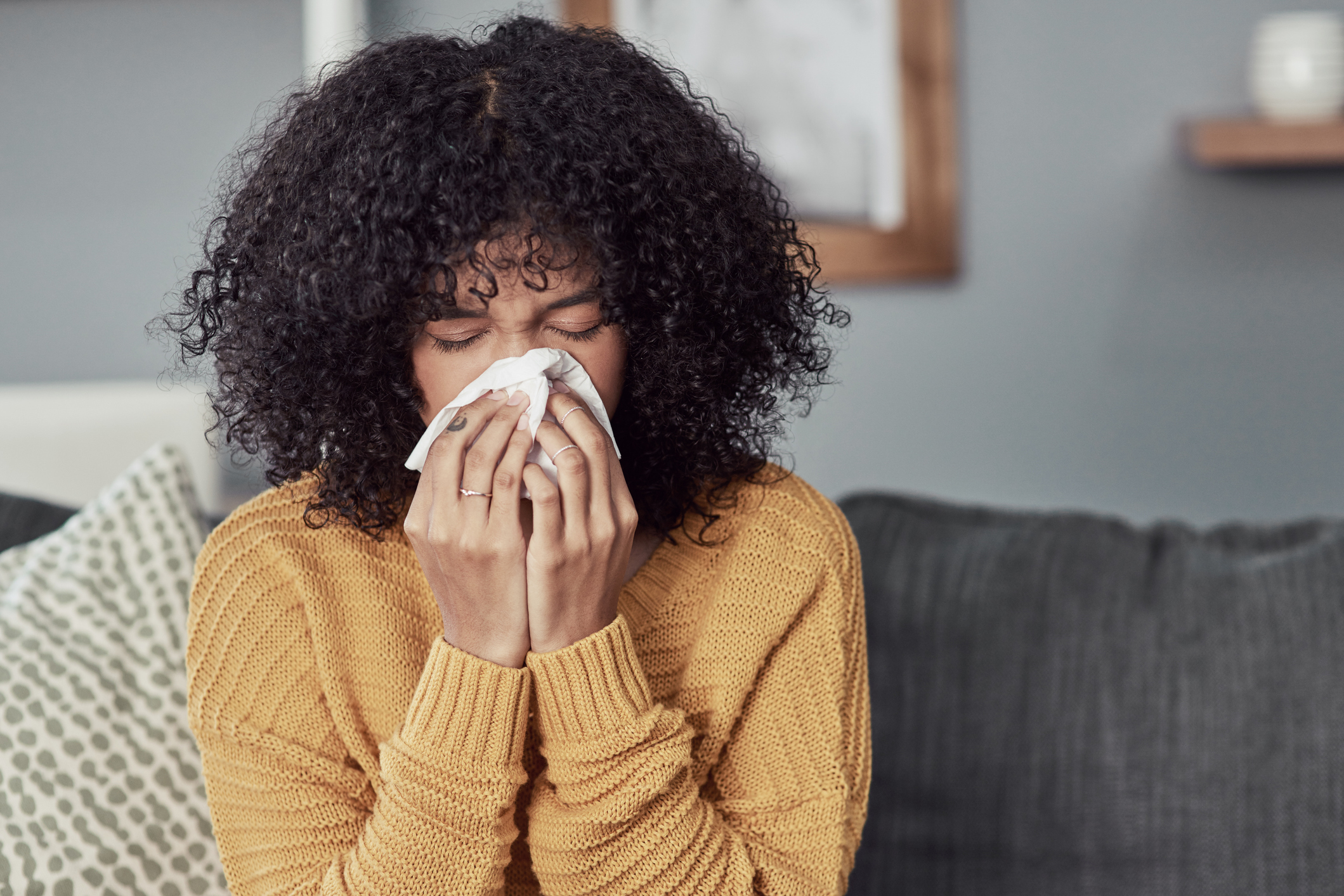 Person blowing their nose into a tissue while sitting on a sofa.