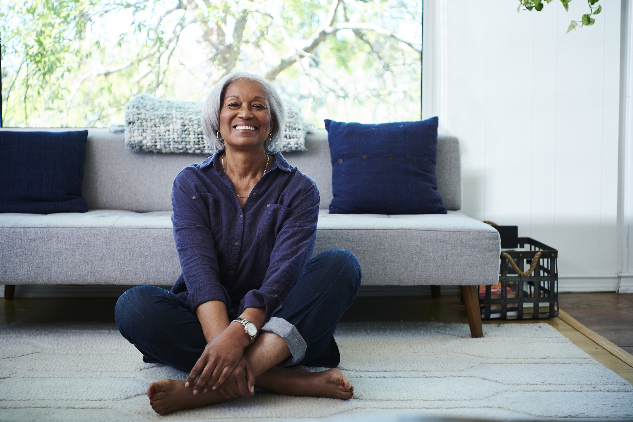 Senior adult smiles while sitting cross-legged on living room floor.
