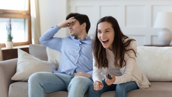 One person hangs their head on a couch while another holds the TV remote with a big smile.