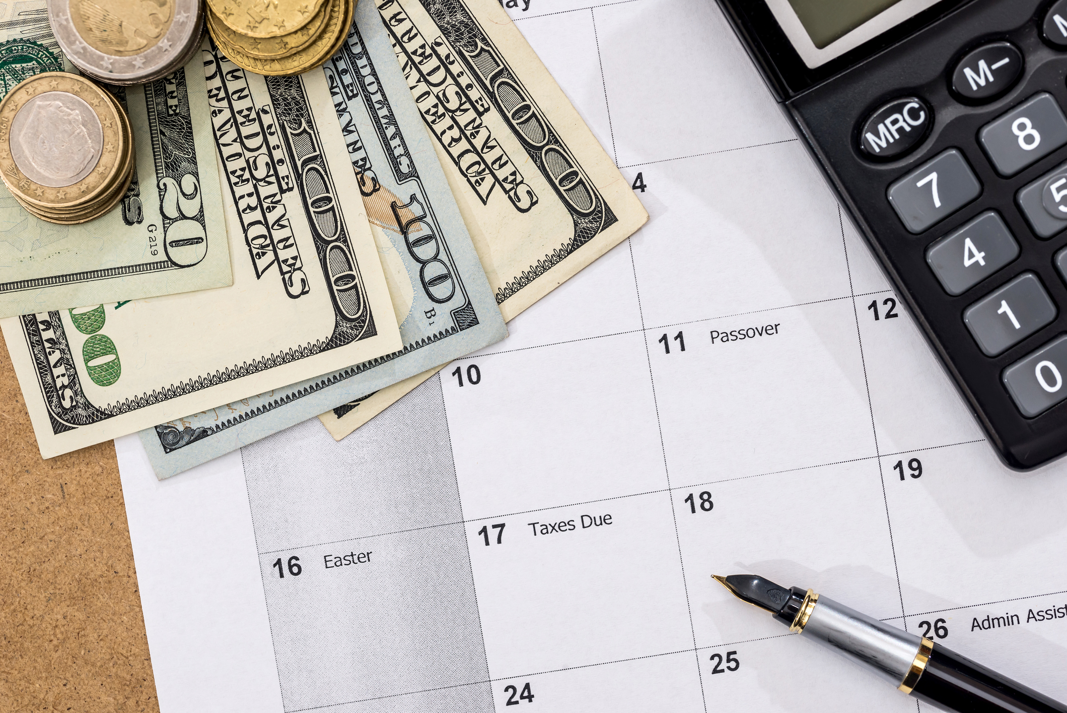Large-denomination dollar bills and various coins resting on a calendar, next to a pen and a calculator.