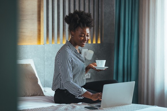 Person sitting on bed with laptop and cup of coffee.