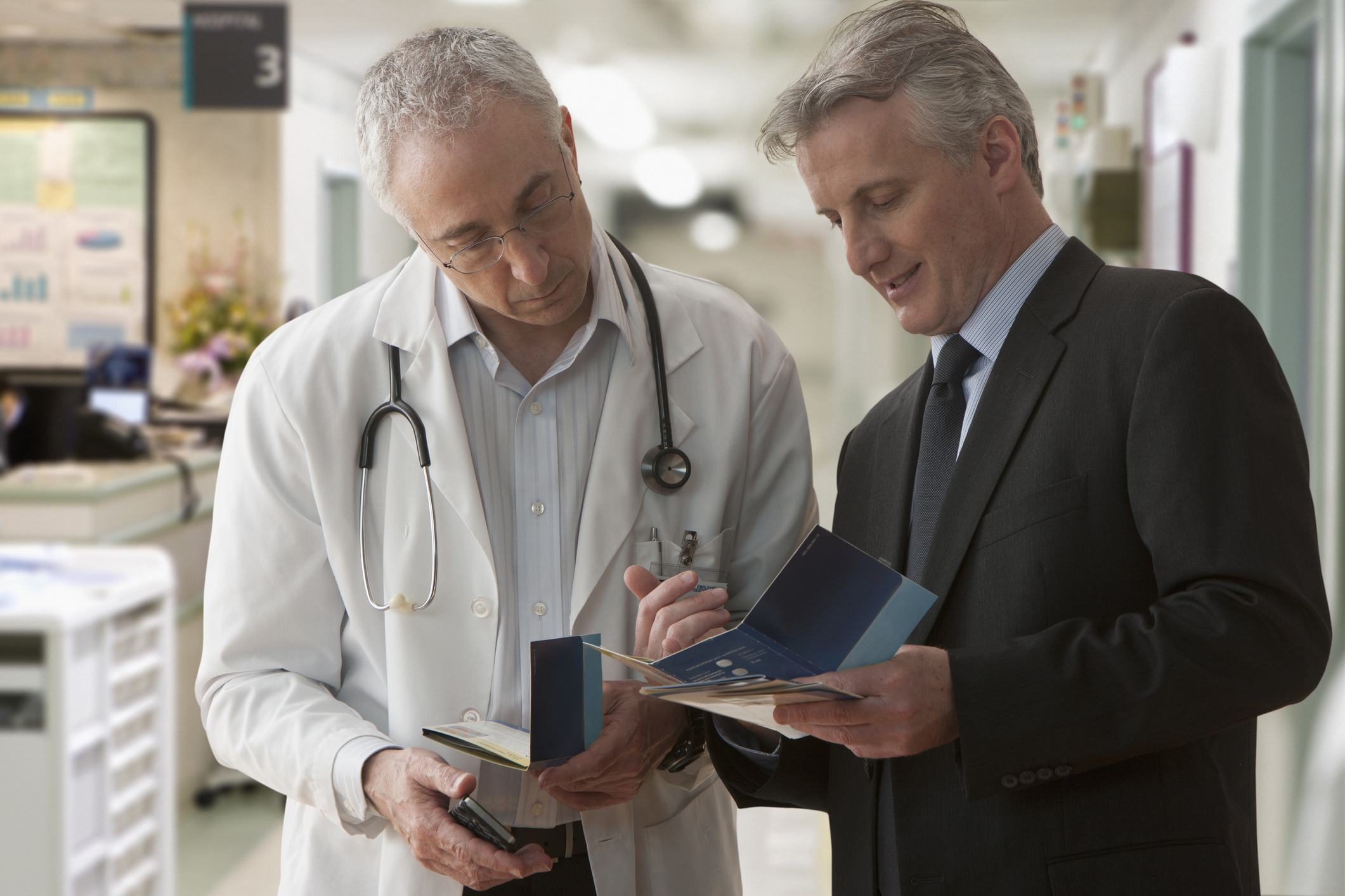 An investor talks with a doctor, gesturing to a pamphlet, while standing in a clinic.