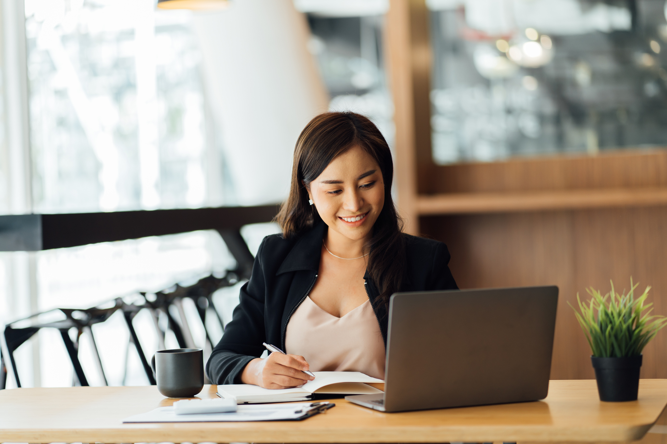 An investor sits at a laptop in a cafe setting and writes in a notebook.