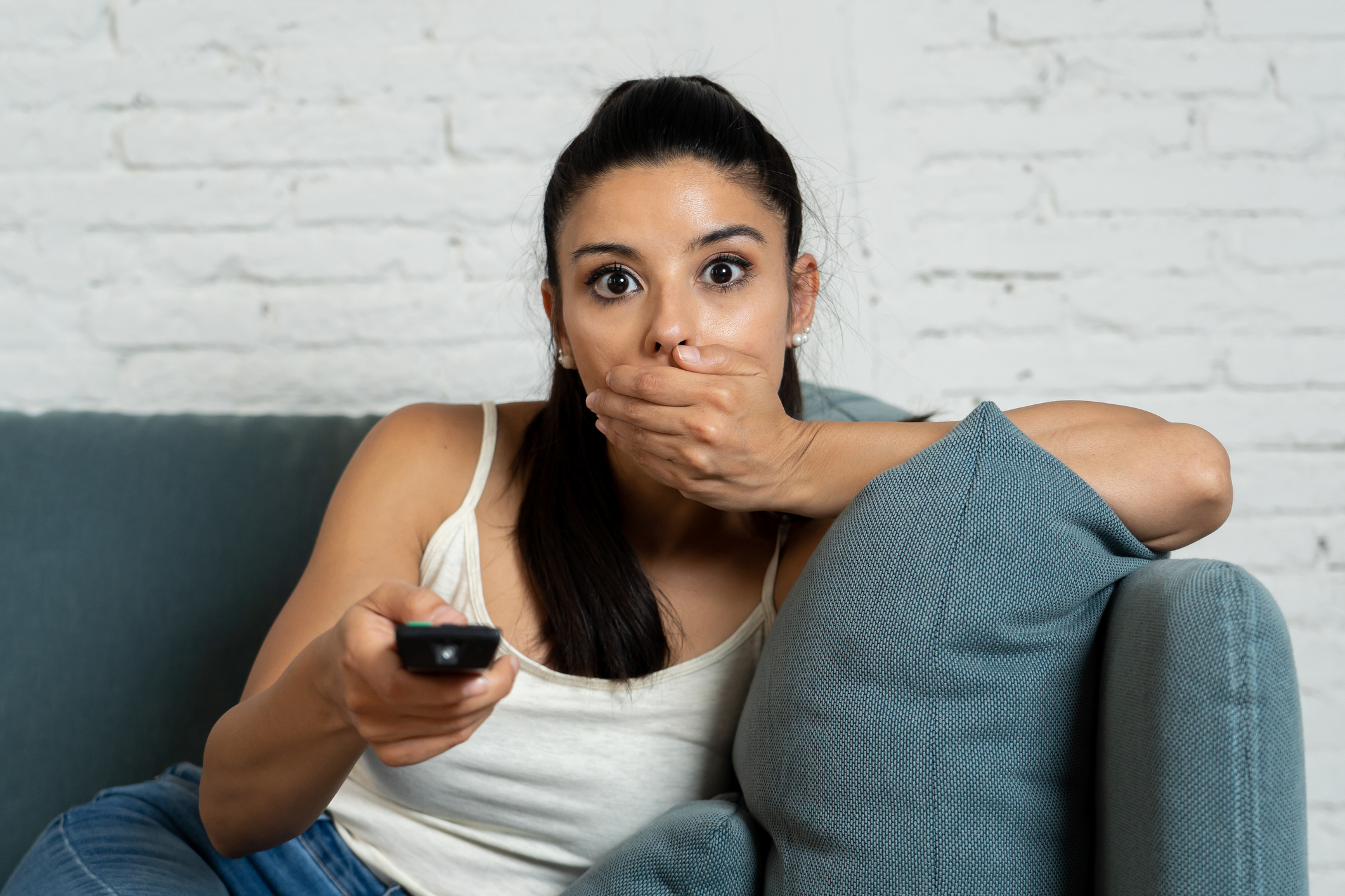 Wide-eyed and frowning person with one hand over their mouth and the other holding a TV remote.