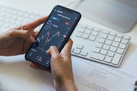 A person holding a smartphone that's displaying a volatile crypto chart with buy and sell buttons above it.