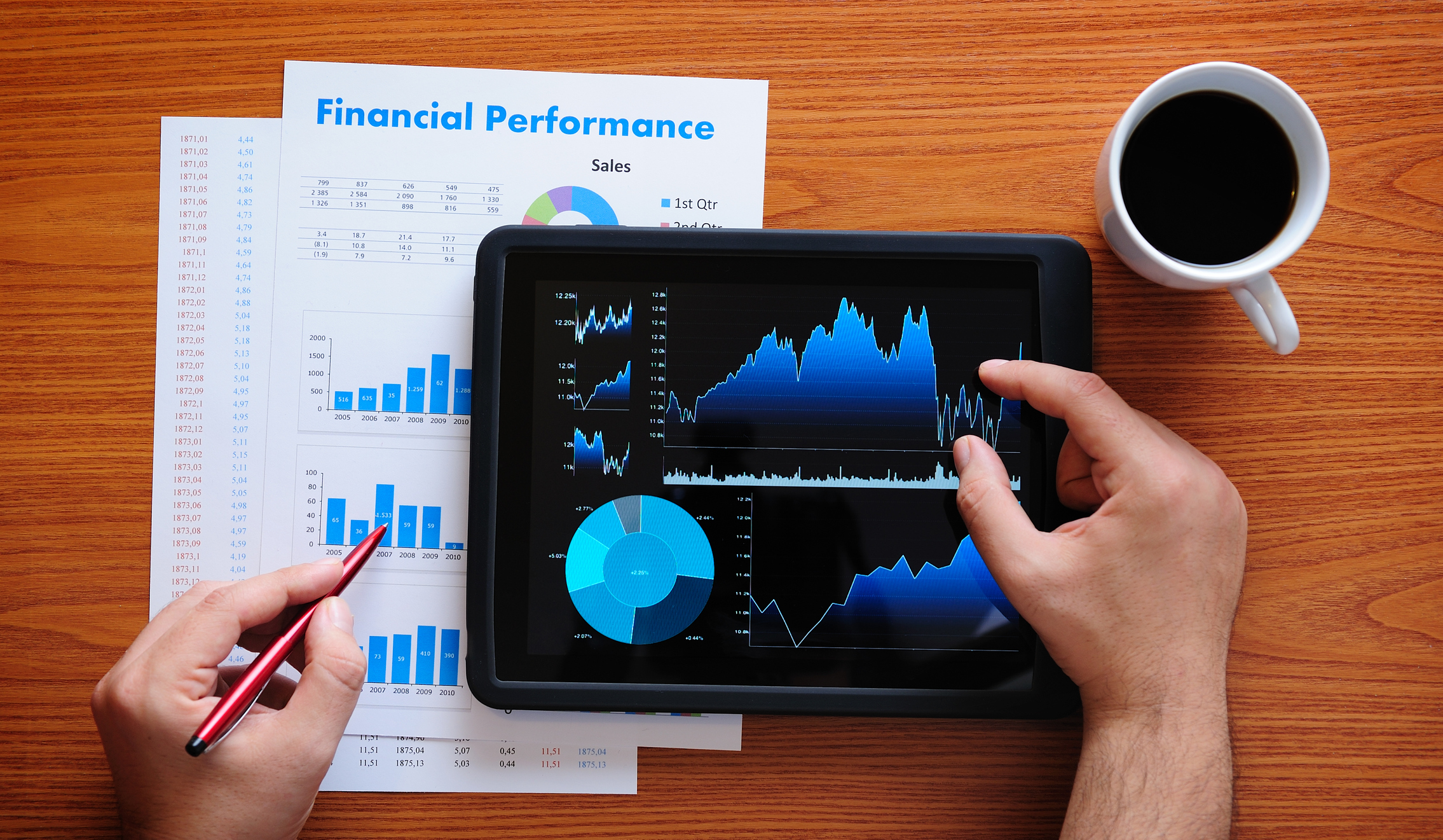 An investors hands hover over paper-based documents and a digital tablet, both of which show financial data.