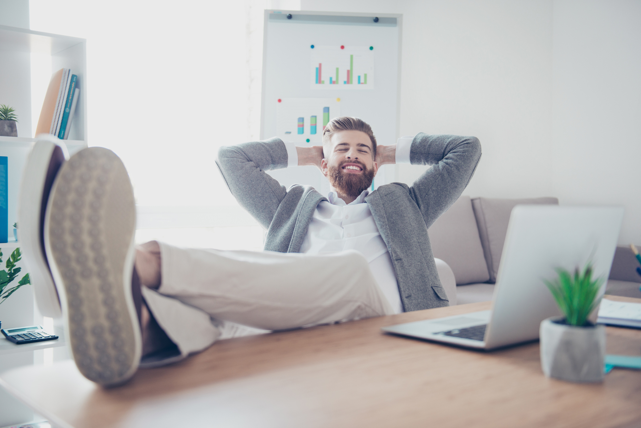 A person has their feet kicked up on a desk, their arms behind their head, and a smile on their face.