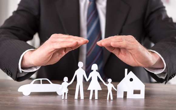 An insurance agent protectively placing their hands above paper cutouts of a house, car, and family of four. 