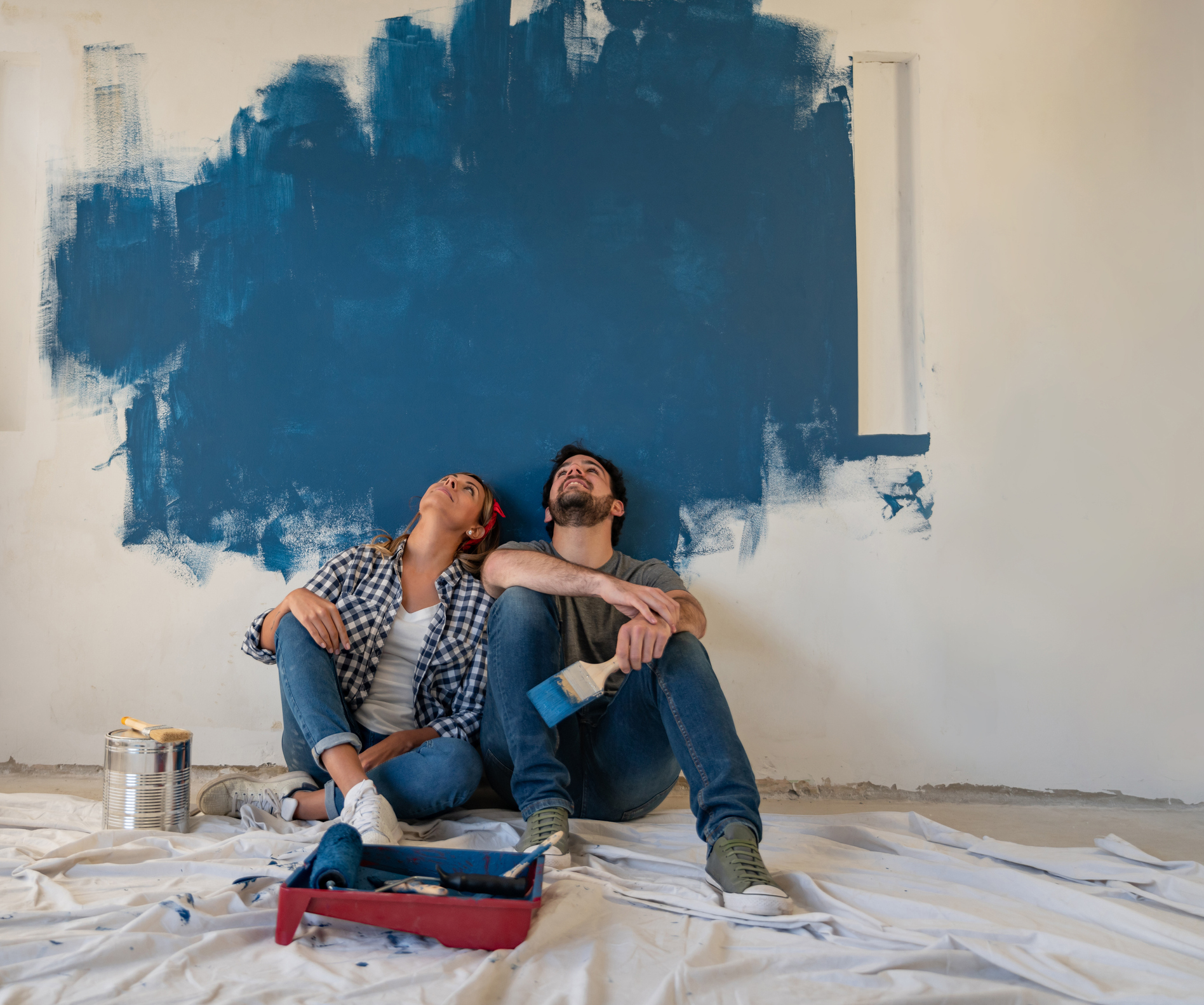 A man and woman sit against a wall while looking up at a blob of blue paint that sporadically coats the wall.