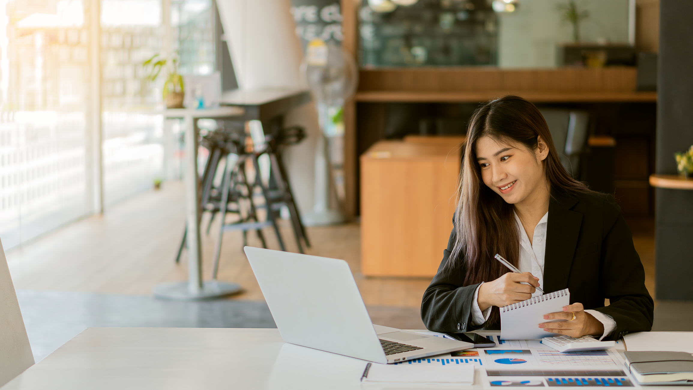 A businessperson smiles while taking notes from information on paper and laptop.