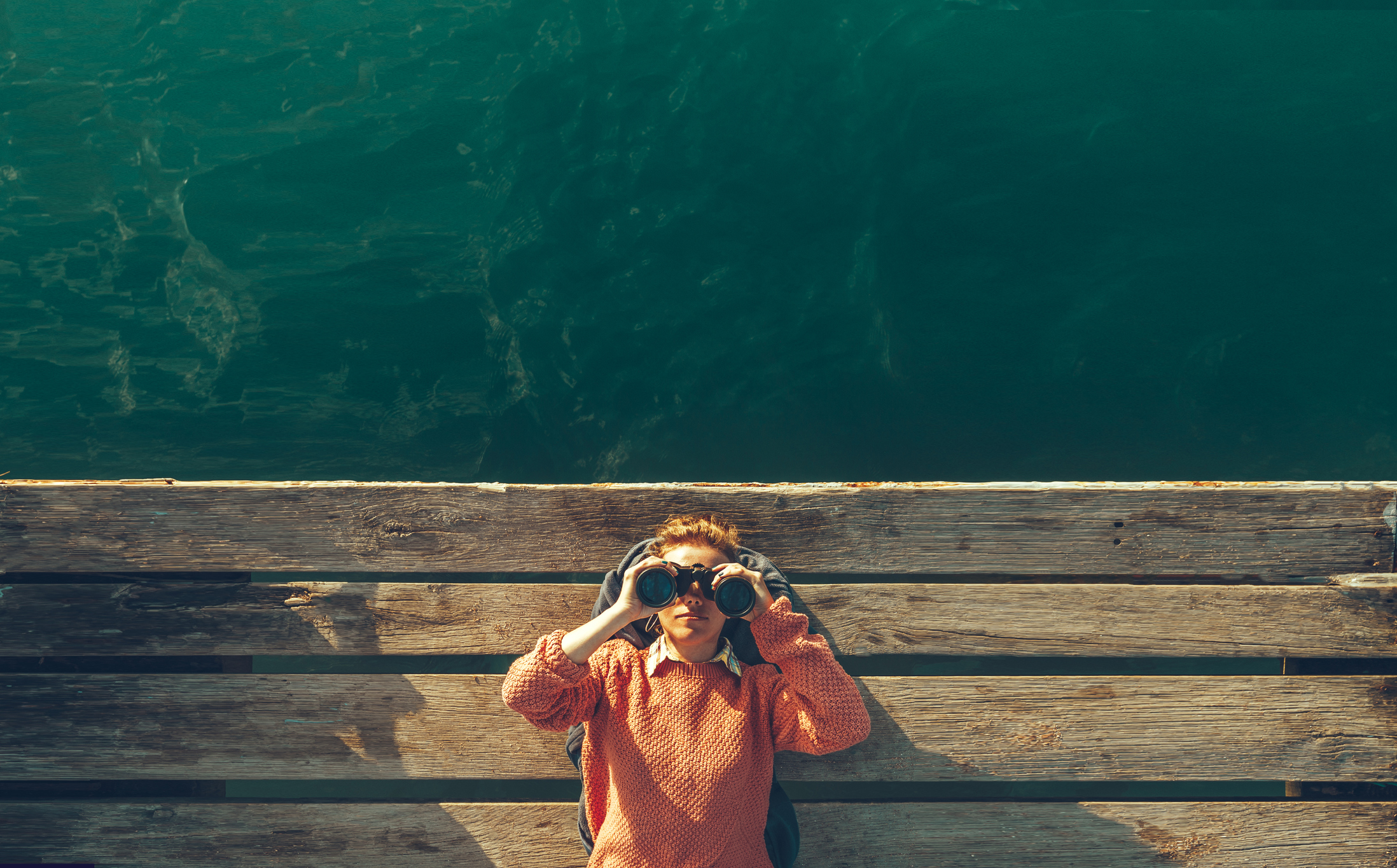 Person lying down on a dock with binoculars looking up.